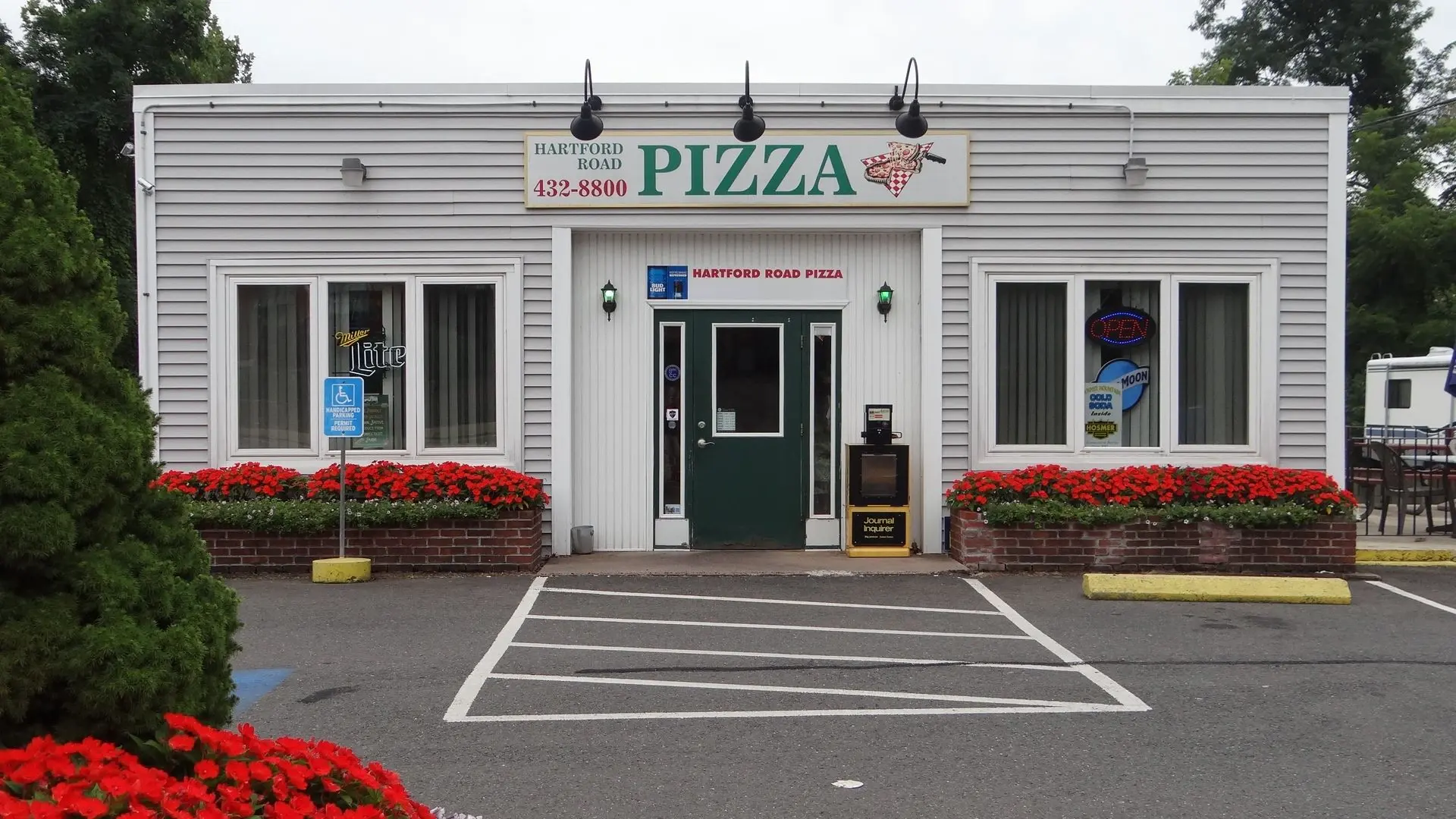 Hartford Road Pizza storefront in daylight, a white clapboard building with a green door and red flower boxes along the brick planters